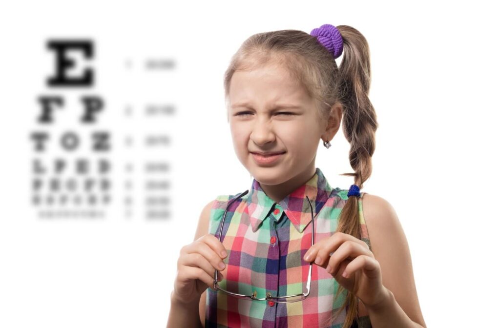 A young girl in front of a Snellen chart squinting her eyes because of myopia and holding glasses in front of her chest.