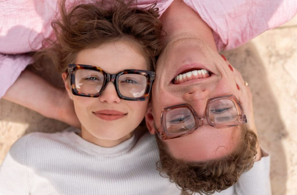 Two smiling individuals wearing thick-framed glasses lie beside each other, one right side up and one upside down