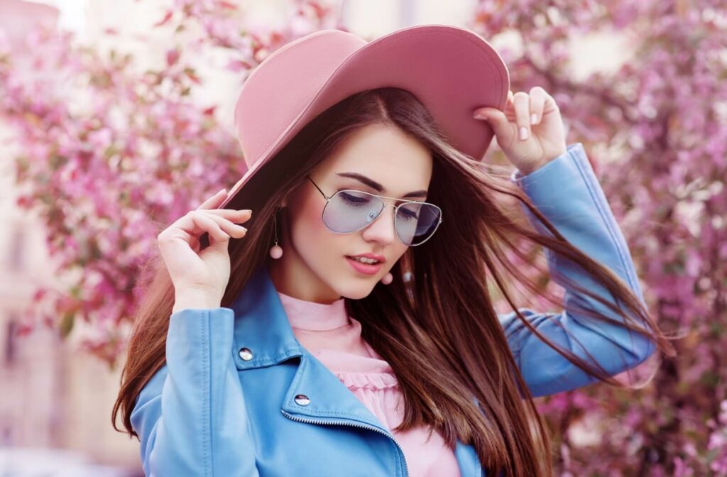 A model in front of cherry blossoms is dressed in blue aviator glasses, a blue jacket, a pink blouse, and a pink hat
