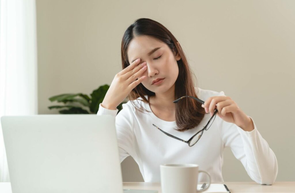 A young adult working from home taking off their glasses to rub their eyes in discomfort from dry eye and floaters.