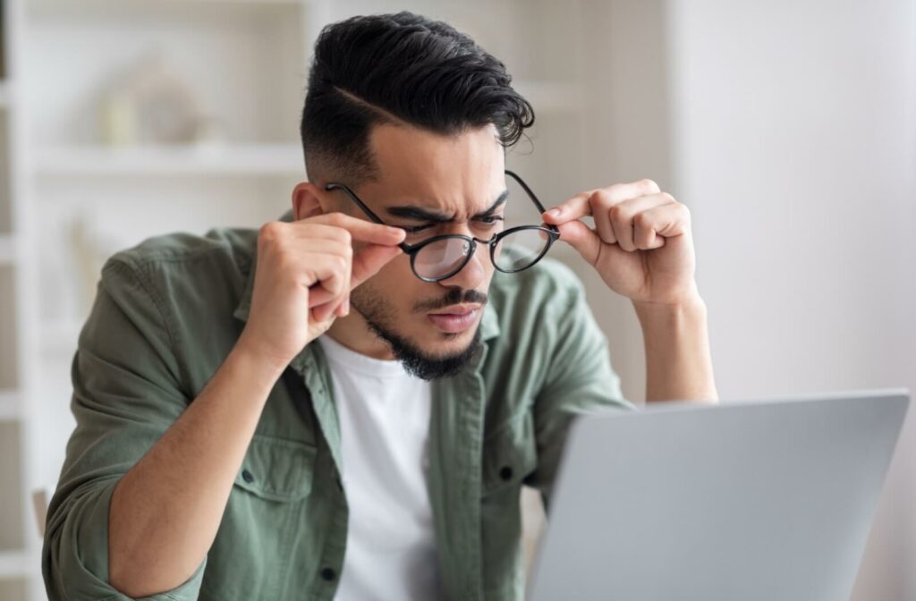 A person holds their glasses and squints at their computer screen, struggling to see.