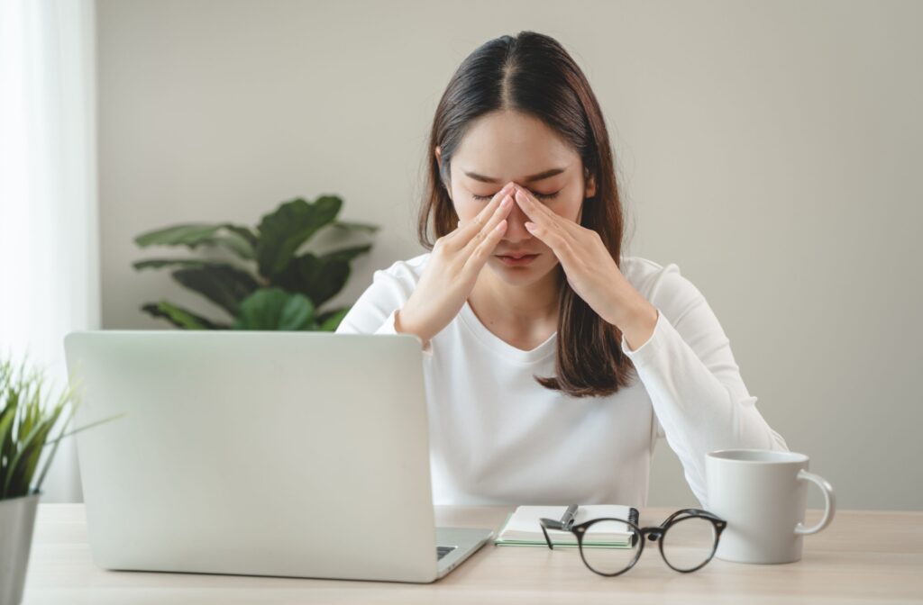 A person sits in front of a computer with their glasses on the table and they rub their dry eyes