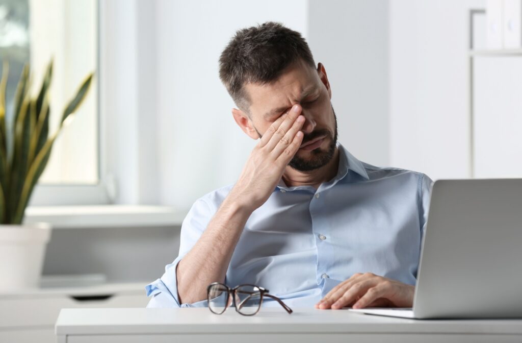 A person sitting at a desk with their glasses off as they rub their irritated eyes