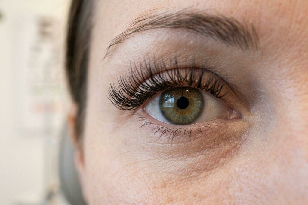 Close-up of a person's open eye with a green-hazel iris, long curled lashes, and a dark brown eyebrow visible above.
